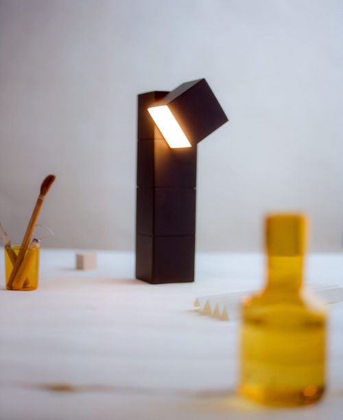 Modern desk lamp made of stacked black cubes with a warm light emanating from the top cube. Yellow containers and white table.