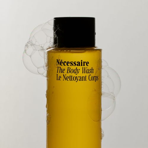 Bottle of Necessaire The Body Wash, yellow liquid with black cap, surrounded by soap bubbles against a light gray background.
