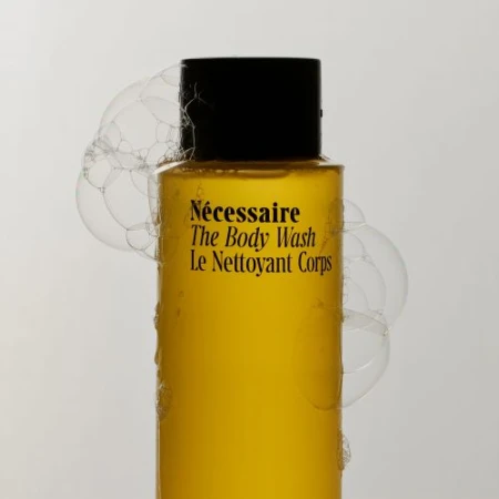 Bottle of Necessaire The Body Wash, yellow liquid with black cap, surrounded by soap bubbles against a light gray background.