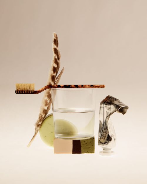 Still life arrangement with toothbrush, feather, glass of water, soap, and a tube of cream against a creamy background.