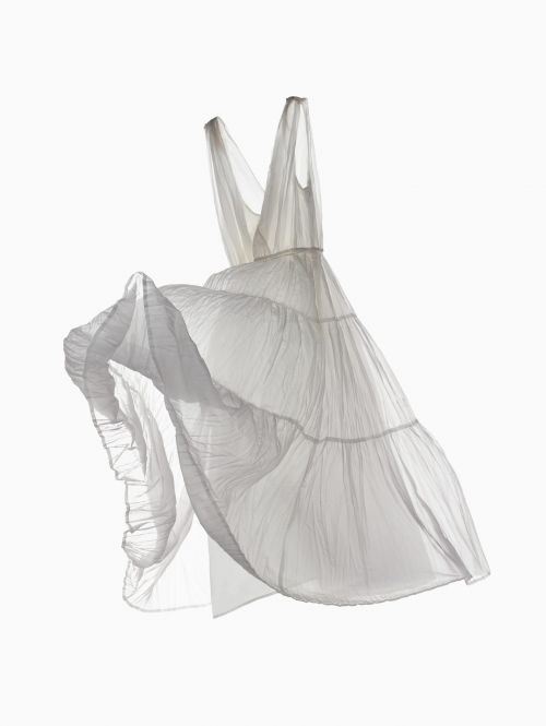 Elegant, sleeveless, semi-sheer white dress with tiered layers is flowing in a bright, clean, minimalist studio setting. Fashion shoot.
