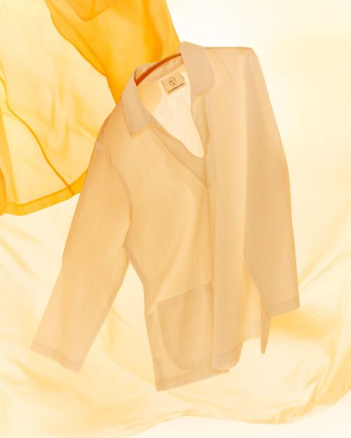 Semi-sheer blouse in white on a hanger, styled with flowing yellow fabric background, studio lighting, and soft, pastel color palette.