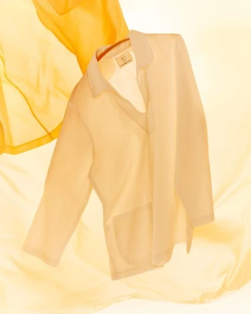 Semi-sheer blouse in white on a hanger, styled with flowing yellow fabric background, studio lighting, and soft, pastel color palette.