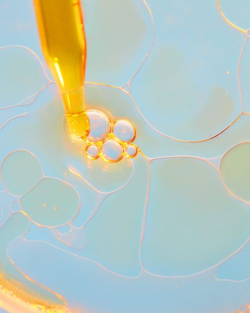 Close-up of golden liquid dropping from pipette into clear fluid with abstract white and blue shapes, creating a vibrant, textured pattern.