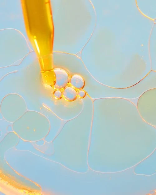 Close-up of golden liquid dropping from pipette into clear fluid with abstract white and blue shapes, creating a vibrant, textured pattern.