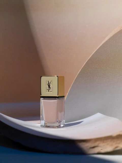 Close-up of a clear nail polish bottle with a gold cap featuring the YSL logo, presented against a light peach and white background.