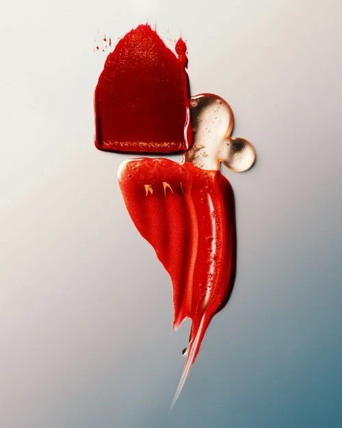 A visually striking image showcasing smears of red lipstick and clear gloss on a gradient white to blue background, highlighting texture.