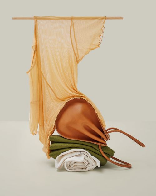 Still life fashion shoot of a draped yellow top, a brown leather purse and folded white and green textiles arranged on a flat surface.