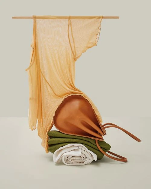 Still life fashion shoot of a draped yellow top, a brown leather purse and folded white and green textiles arranged on a flat surface.