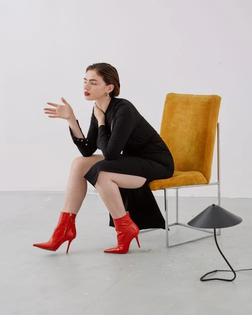A woman in a black dress and red ankle boots sits on a yellow chair in a bright, minimal studio with a black lamp nearby.