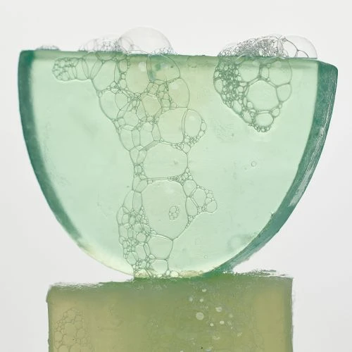 Close-up of a translucent green soap bar with bubbles on top, set against a plain white background, highlighting its texture.