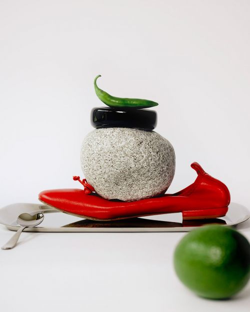 Still life: red shoe, stone, black ring, green chili, lime on silver tray against white background. Editorial food and fashion.