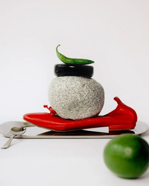 Still life: red shoe, stone, black ring, green chili, lime on silver tray against white background. Editorial food and fashion.