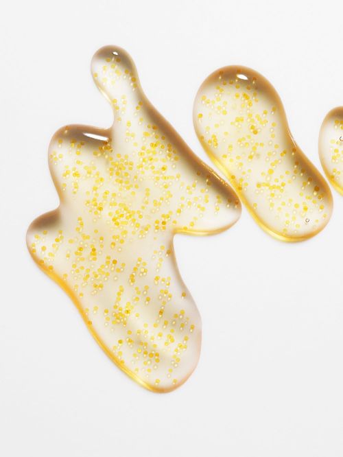 Macro photograph featuring three clear gel blobs, each filled with suspended golden particles, on a bright white surface. Beauty shot.