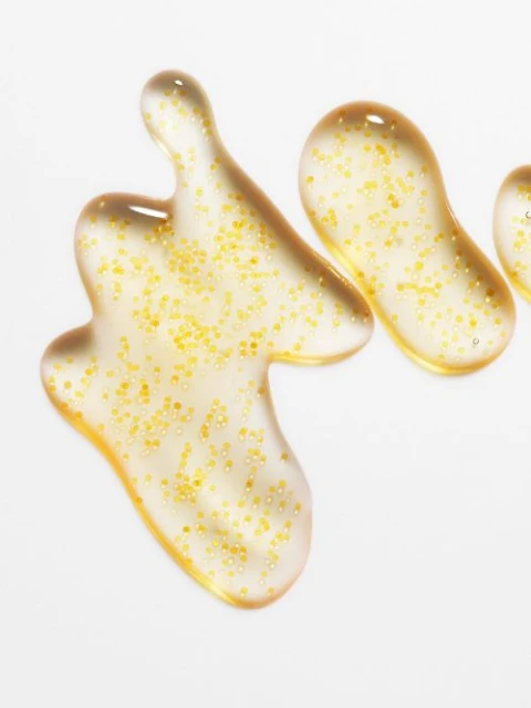 Macro photograph featuring three clear gel blobs, each filled with suspended golden particles, on a bright white surface. Beauty shot.