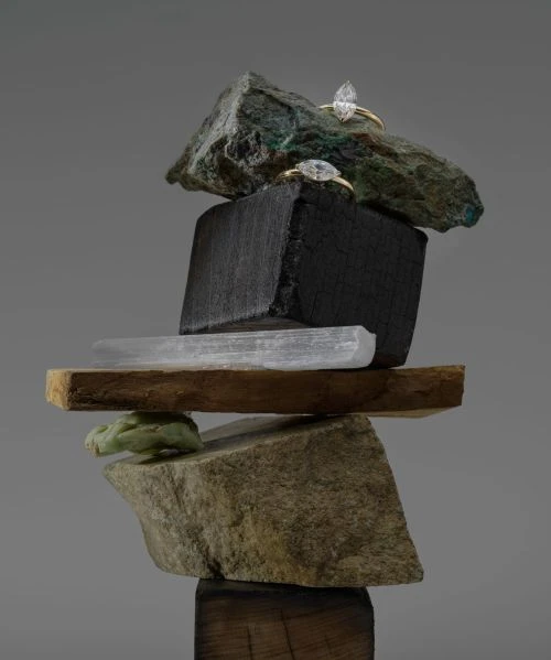 Two engagement rings are balanced on a sculpture made of stones, wood, and minerals against a grey background, showcasing jewelry design.