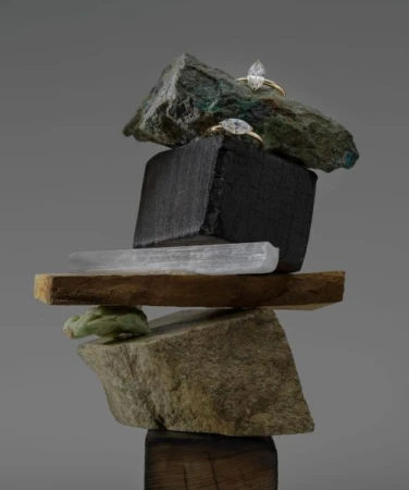Two engagement rings are balanced on a sculpture made of stones, wood, and minerals against a grey background, showcasing jewelry design.