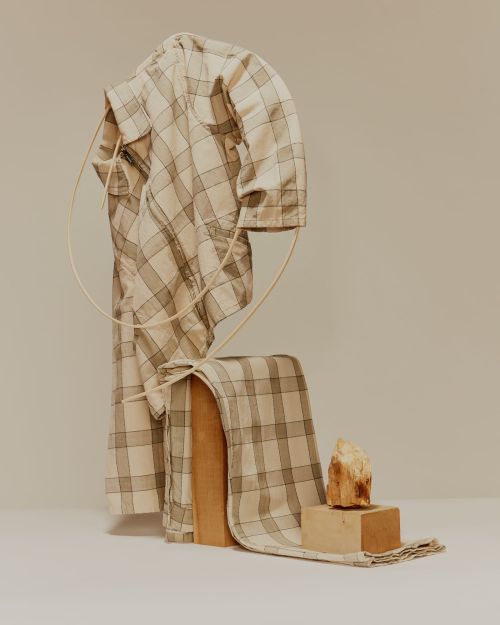 Still life of a folded, checkered shirt draped on a wood block, next to a small stone on a block, against an off-white backdrop.