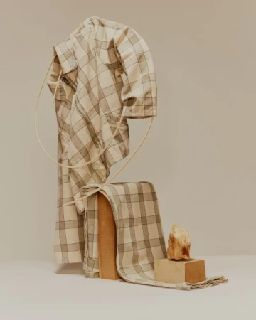 Still life of a folded, checkered shirt draped on a wood block, next to a small stone on a block, against an off-white backdrop.