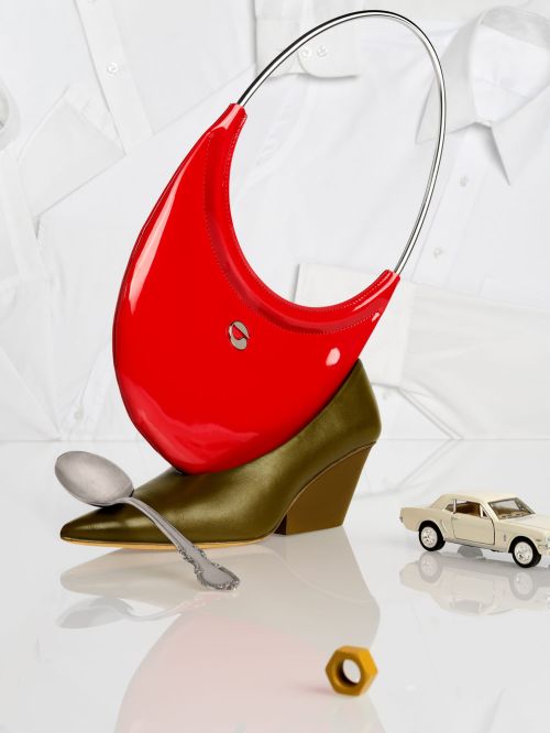 Still life with a red purse resting on a heel, accented by a spoon, a car toy, and a golden nut against a white shirt backdrop.
