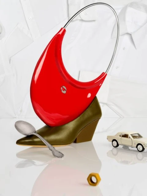 Still life with a red purse resting on a heel, accented by a spoon, a car toy, and a golden nut against a white shirt backdrop.