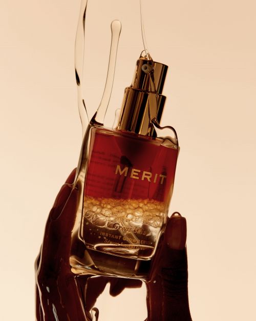 Close-up of a person holding a bottle of Merit skincare serum with a liquid pouring over it against a neutral background.