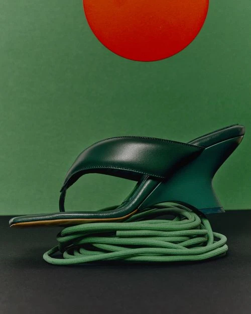 Elevated studio shot of a single green sandal with a geometric heel perched atop bundled green cord against a two-tone green backdrop.