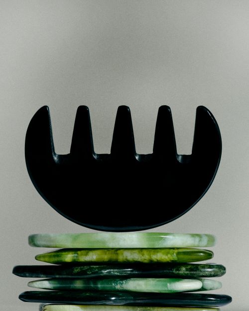 Black guasha comb atop a stack of translucent green stone bracelets on a neutral backdrop, showcasing beauty and self-care.