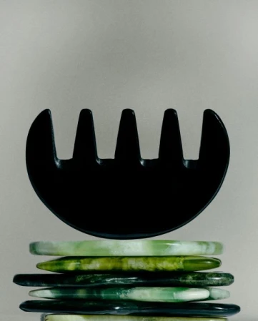 Black guasha comb atop a stack of translucent green stone bracelets on a neutral backdrop, showcasing beauty and self-care.