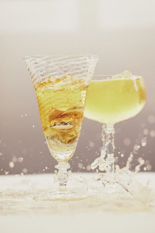Two clear stemmed glasses with gold-colored liquid. One glass spills liquid creating a splash. Set against a gradient background.