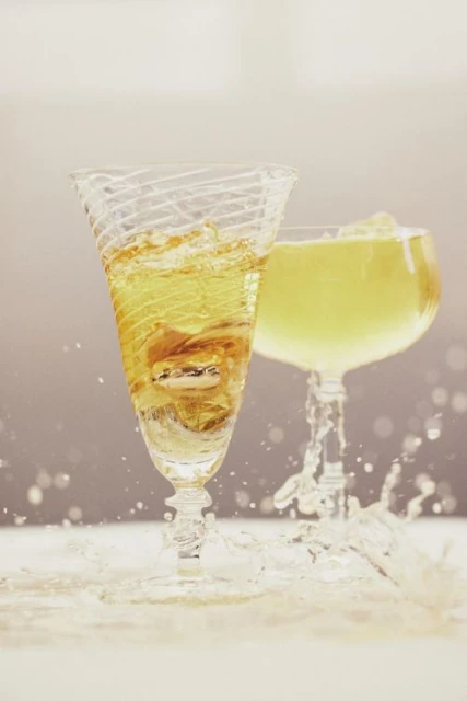 Two clear stemmed glasses with gold-colored liquid. One glass spills liquid creating a splash. Set against a gradient background.