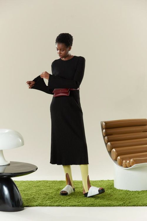 Elegant Black Woman in Ribbed Midi Dress with Burgundy Belt Bag Stands on Green Rug in Fashion Editorial Photo.