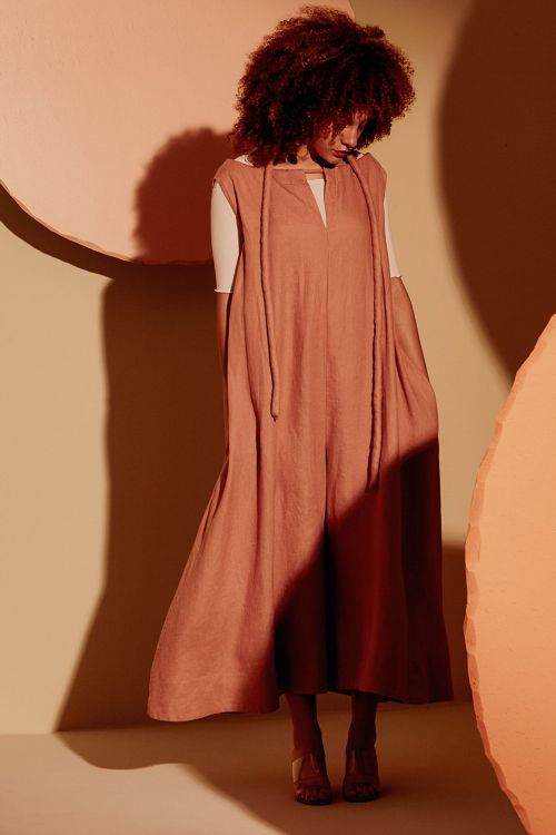 Studio shot of a woman with red curly hair wearing a peach jumpsuit and clear heeled sandals against peach and beige backdrop.