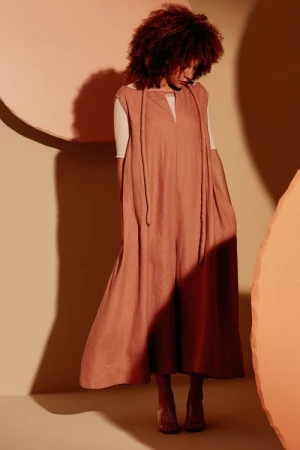 Studio shot of a woman with red curly hair wearing a peach jumpsuit and clear heeled sandals against peach and beige backdrop.