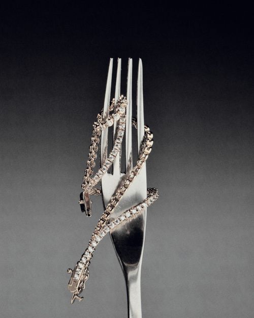Diamond tennis bracelet coiled around a silver fork against a grey gradient backdrop, emphasizing luxury and culinary fusion in jewelry.