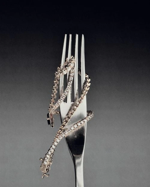 Diamond tennis bracelet coiled around a silver fork against a grey gradient backdrop, emphasizing luxury and culinary fusion in jewelry.
