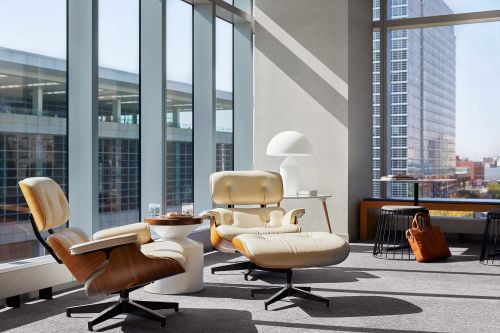 Modern interior with Eames lounge chairs and ottoman near large windows with a cityscape view, bathed in natural light.