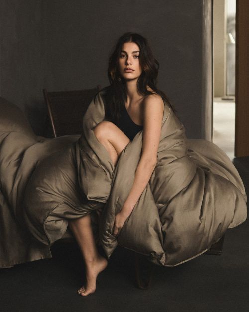 Woman wrapped in a blanket sitting on a bed inside an indoor space, looking at the camera with a neutral facial expression. 