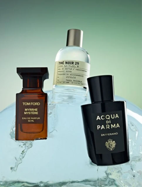 Three perfume bottles by Tom Ford, Le Labo, and Acqua di Parma are displayed on an icy surface with a green and white gradient background.