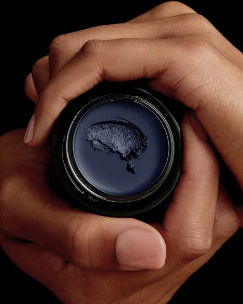 Close-up shot of hands holding a jar of dark blue cream against a black background, showcasing skincare or cosmetic product.