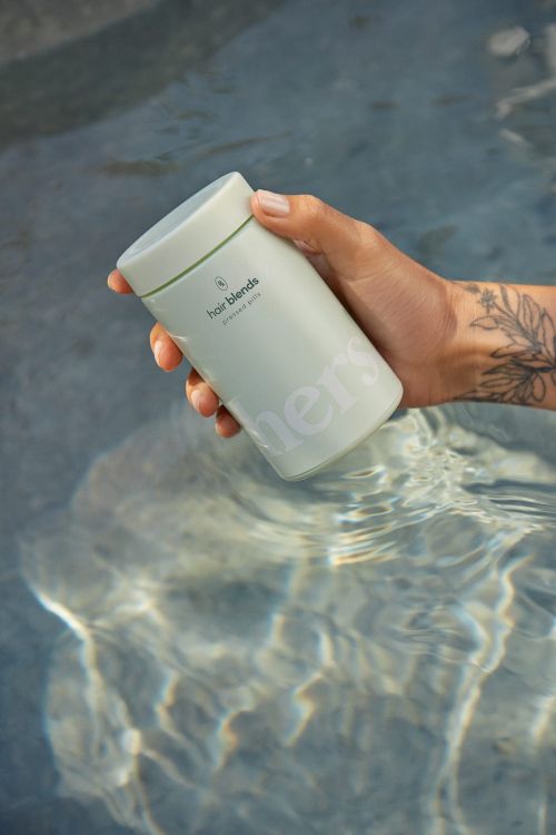 Person holds light-green hair product in water with sunlight reflecting. Product has brand text and simple geometric logo.