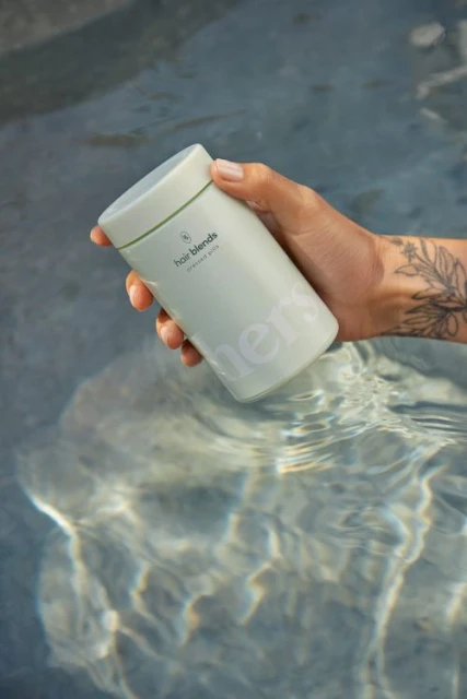 Person holds light-green hair product in water with sunlight reflecting. Product has brand text and simple geometric logo.