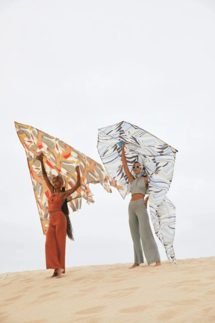 Two women in matching outfits holding patterned scarves on a sandy dune, posing in front of a bright, overcast sky in an outdoor setting.