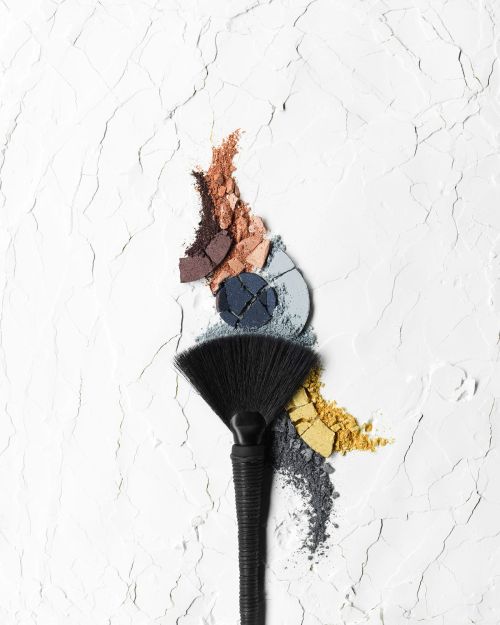 Overhead studio shot of a makeup brush with colorful eyeshadow powders scattered on a textured white background, creating a vibrant beauty image.