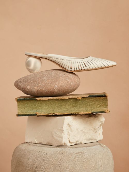 Studio shot of a white heeled shoe balanced on a rock, book, and white block, stacked on a pedestal against a peach background.