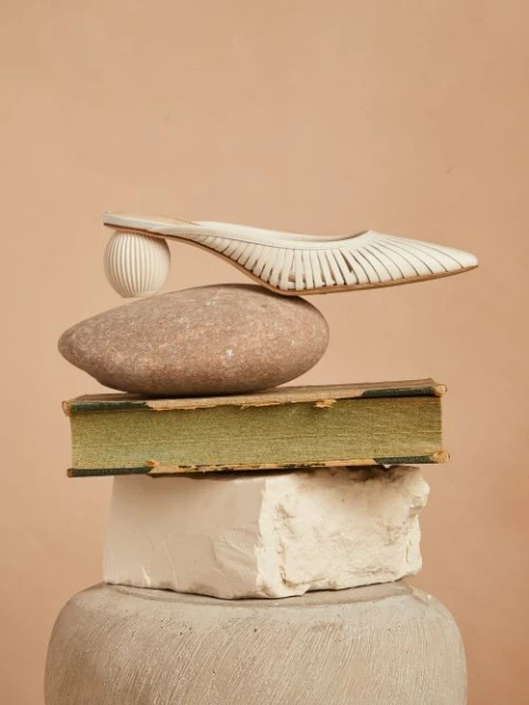 Studio shot of a white heeled shoe balanced on a rock, book, and white block, stacked on a pedestal against a peach background.