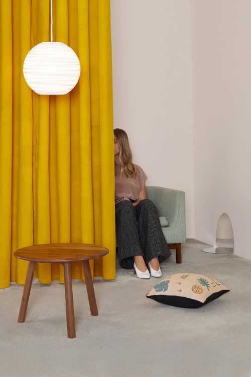Interior scene featuring a partially visible woman sitting in a chair behind a yellow curtain, with furniture and decor elements.