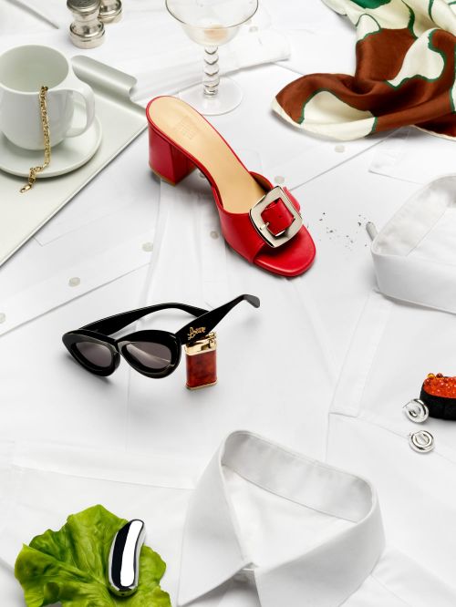 Flat lay of red heeled mule, sunglasses, scarf, cuff links, cup, lighter, and jewelry on white shirts, creating a fashion still life.