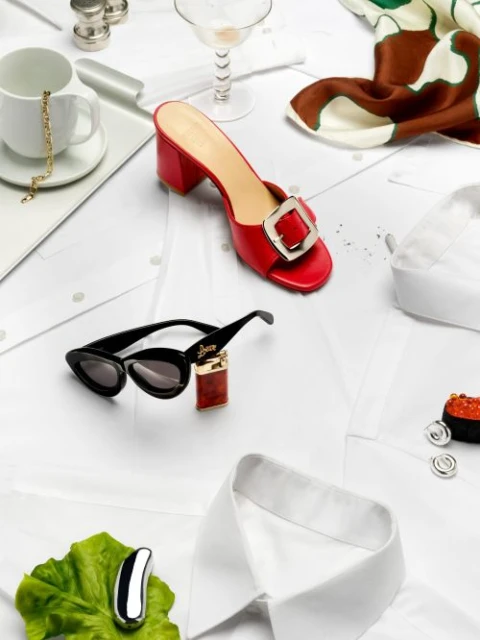Flat lay of red heeled mule, sunglasses, scarf, cuff links, cup, lighter, and jewelry on white shirts, creating a fashion still life.