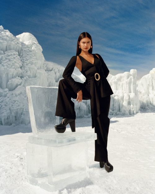 A woman in a black pantsuit poses in a snowy landscape next to a chair made of ice, with snow and a blue sky with white clouds in the backdrop.